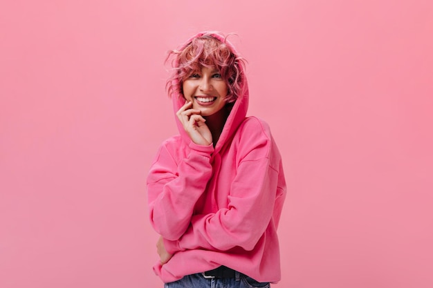 Heureuse jeune femme aux cheveux roses dans un sweat à capuche fuchsia surdimensionné lumineux sourit largement et se penche sur la caméra sur fond isolé