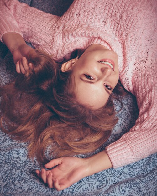 Heureuse jeune femme aux cheveux rouges dans un pull rose allongé sur le lit