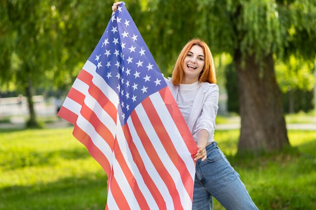 Heureuse jeune femme aux cheveux rouges posant avec le drapeau national des États-Unis debout à l'extérieur dans le parc d'été. Fille positive célébrant la fête de l'indépendance des États-Unis.