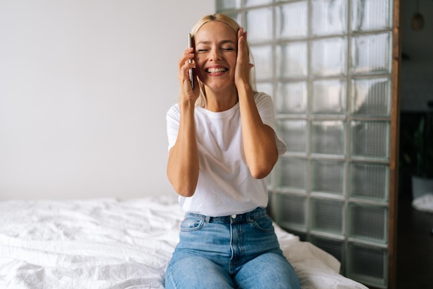 Heureuse jeune femme blonde aux yeux fermés s'asseoir sur le lit à l'aide d'un téléphone portable a obtenu une offre fantastique lire de bonnes nouvelles se sent excitée appel tant attendu