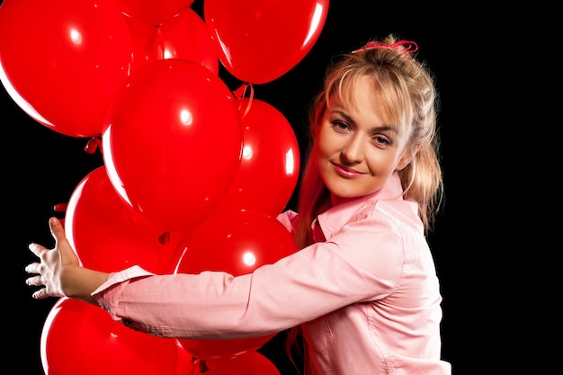 Heureuse jeune femme blonde en blouse rose tenant des ballons d'hélium rouges. Isolé sur fond noir