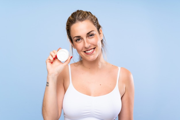 Heureuse jeune femme blonde avec un hydratant sur un mur bleu isolé