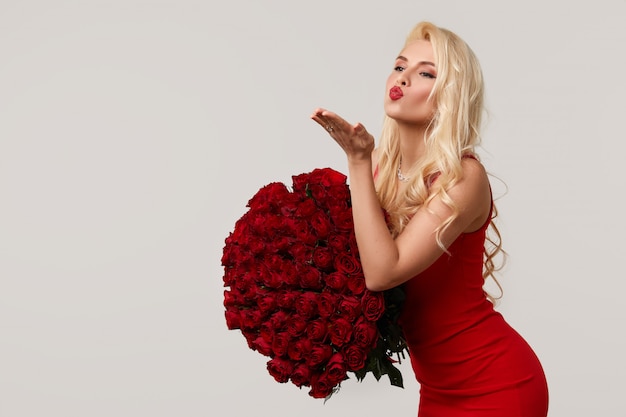 Heureuse jeune femme blonde tenant un grand bouquet de roses rouges en cadeau pour le 8 mars ou la Saint-Valentin. Elle souffle un baiser. Fond gris neutre