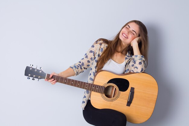 Heureuse jeune femme en blouse beige et pantalon noir tenant une guitare sur fond gris en studio