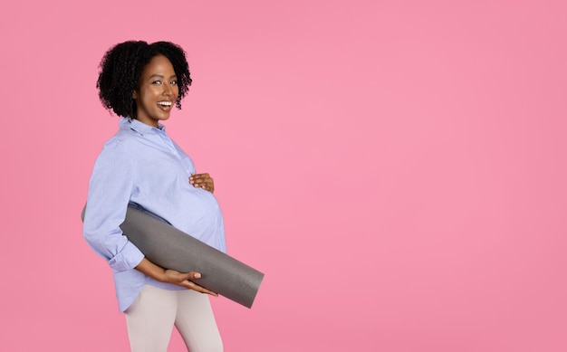 Heureuse jeune femme bouclée afro-américaine enceinte en tenue décontractée avec gros tapis de maintien du ventre