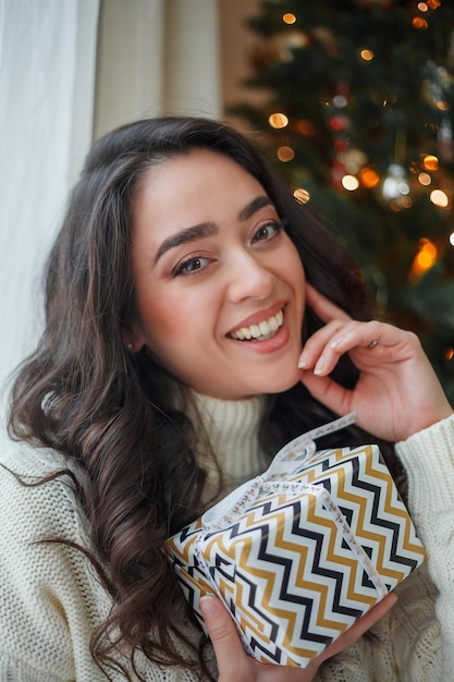 Heureuse jeune femme bouclée aux cheveux noirs dans un pull tricoté léger prépare des cadeaux près de l'arbre de Noël Un modèle élégant fait des achats en ligne pendant les vacances d'hiver Nouvel An