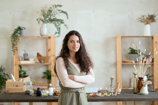 Heureuse jeune femme brune croisant les bras par la poitrine en se tenant debout contre le lieu de travail avec des ingrédients pour la fabrication de produits cosmétiques naturels