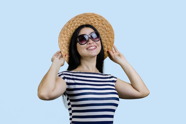 Heureuse jeune femme brune gaie posant dans un chapeau de paille et des lunettes de soleil isolées sur le backgro bleu pâle