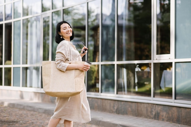 Heureuse jeune femme brune avec des lunettes de soleil et un sac tenant un café marchant dans la ville. Portrait de mode de vie d'une femme souriante