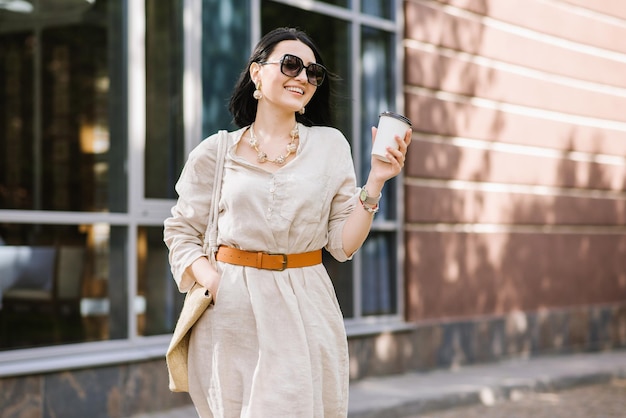 Heureuse jeune femme brune avec des lunettes de soleil et un sac tenant un café marchant dans la ville. Portrait de mode de vie d'une femme souriante