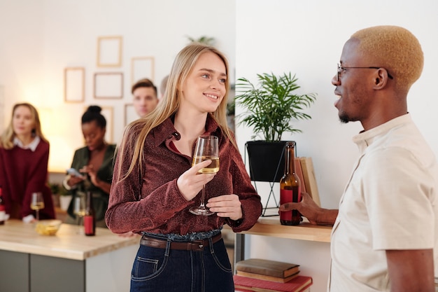 Heureuse jeune femme brune en pull rose flûte de champagne tintant avec celle de son petit ami tout en profitant d'une fête à la maison avec des amis