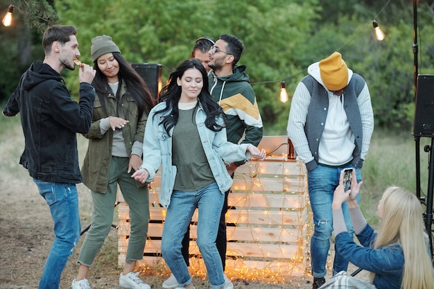 Heureuse jeune femme brune en tenue décontractée dansant et s'amusant parmi ses amis le week-end dans un environnement naturel