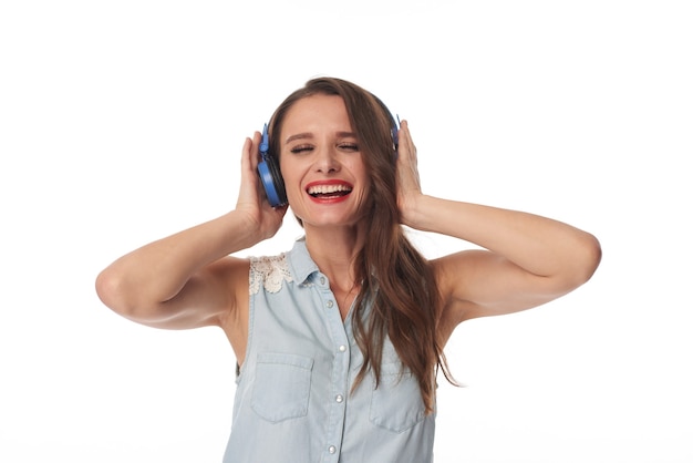 Heureuse jeune femme caucasienne aux yeux fermés tenant des écouteurs bleus tout en écoutant de la musique énergique, fond isolé