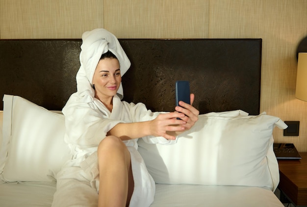 Heureuse jeune femme caucasienne en peignoir blanc et tête enveloppée dans une serviette de bain allongée sur le lit dans la chambre de l'hôtel et sourit en regardant la caméra sur son smartphone tout en prenant un autoportrait, selfie