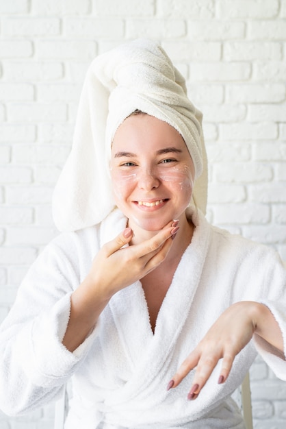 Heureuse jeune femme caucasienne en serviette de bain blanche, appliquer la crème pour le visage à la maison