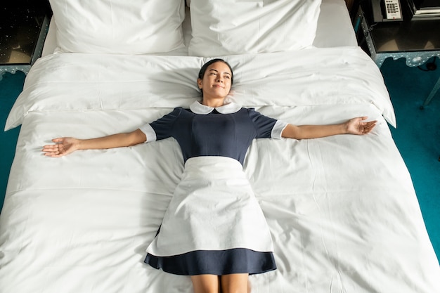Heureuse jeune femme de chambre en uniforme allongée sur un grand lit double