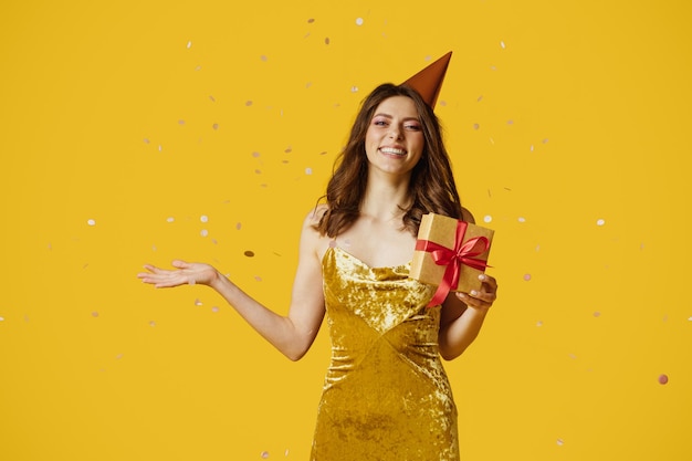 Heureuse jeune femme en chapeau d'anniversaire et robe tenant une boîte-cadeau debout sur fond jaune avec des confettis tombant