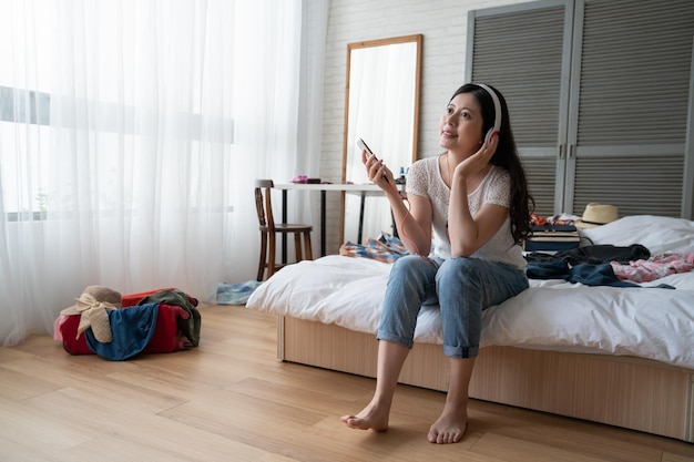 Heureuse jeune femme chinoise asiatique appréciant la musique alors qu'elle se détend au lit avec un téléphone portable et un casque stéréo souriant d'appréciation. adolescente en vacances d'emballage pour le nouveau semestre valise