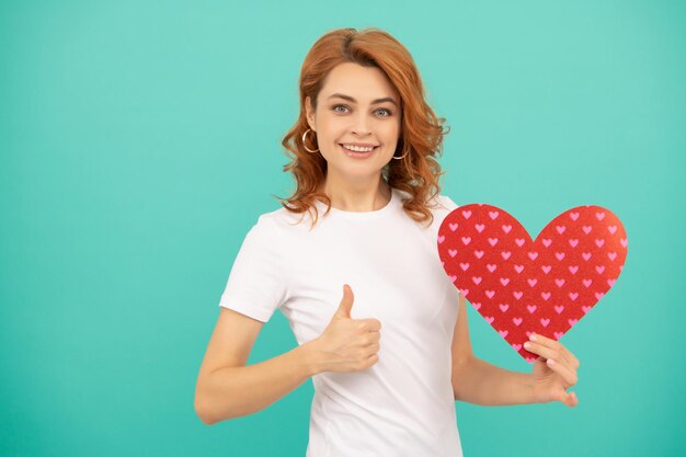 Heureuse jeune femme avec coeur rouge sur fond bleu pouce vers le haut