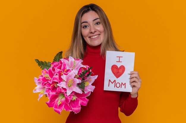 heureuse jeune femme en col roulé rouge tenant une carte de voeux et un bouquet de fleurs. J'aime maman. Fête des mères