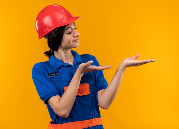 Heureuse jeune femme de construction en uniforme faisant semblant de tenir et de pointer vers quelque chose d'isolé sur un mur jaune avec espace de copie