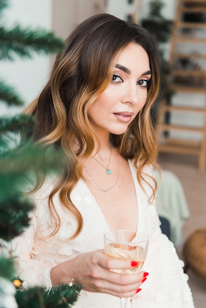 Heureuse jeune femme avec une coupe de champagne posant à l'arbre de Noël en regardant la caméra