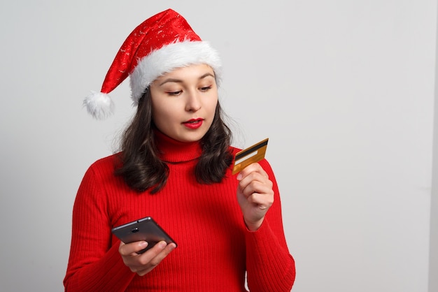 Heureuse jeune femme dans un bonnet de Noël et un pull rouge tient un smartphone et une carte de crédit sur blanc.