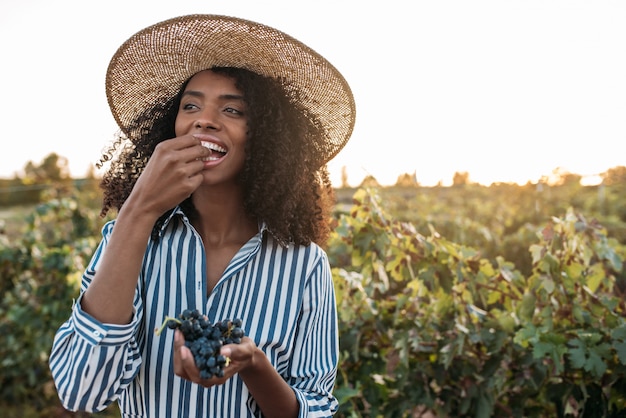 Heureuse jeune femme dans un chapeau de paille, manger des raisins dans un vignoble