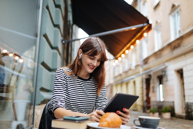 Heureuse jeune femme dans des écouteurs sans fil assis à une table de café et utilisant une tablette numérique