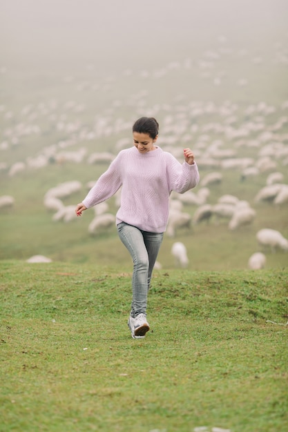 Heureuse jeune femme dans un pull contre des troupeaux de moutons dans les montagnes en jour de brouillard