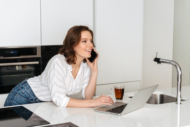 Heureuse jeune femme debout dans la cuisine à la maison, à l'aide de téléphone mobile