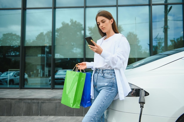 Heureuse jeune femme debout sur le parking de la ville près de la voiture électrique chargeant la batterie de l'automobile depuis la petite station de la ville tenant des sacs à provisions