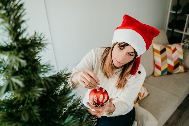 Heureuse jeune femme décorant son arbre de Noël déguisé avec un bonnet de Noel.