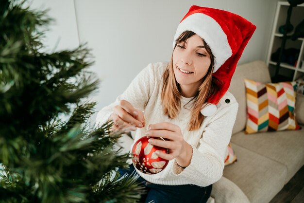 Heureuse jeune femme décorant son arbre de Noël déguisé avec un bonnet de Noel.