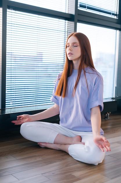 Heureuse jeune femme détendue méditant assise sur le sol dans une pose de lotus par fenêtre dans une salle de bureau lumineuse