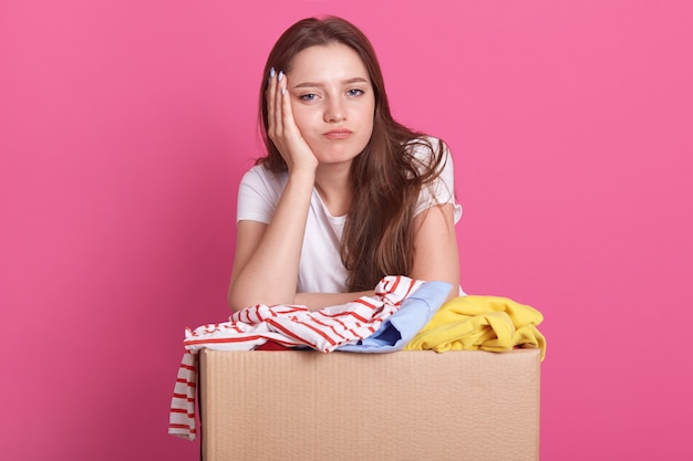 heureuse jeune femme avec don de vêtements debout sur rose, emballant des vêtements réutilisables pour les pauvres, les femmes gardent la main sous le menton, s'ennuyant à la caméra.