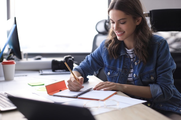 Heureuse jeune femme écrivant quelque chose tout en travaillant au bureau.