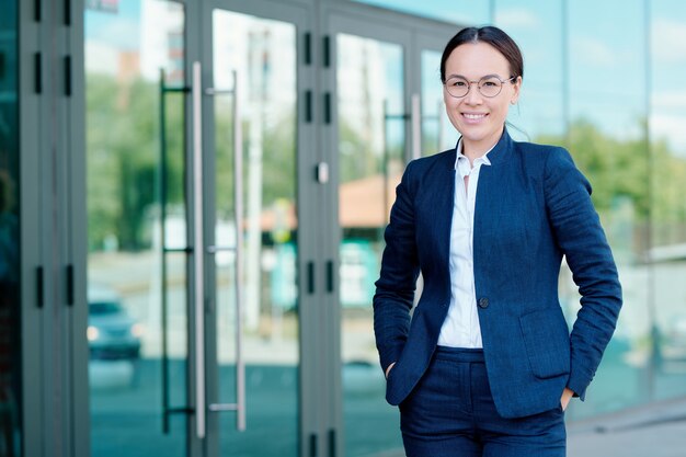 Heureuse jeune femme élégante en tenue de soirée debout devant la caméra sur fond de l'extérieur de l'immeuble de bureaux