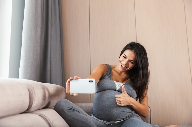 Heureuse jeune femme enceinte assise sur un canapé à la maison, prenant un selfie