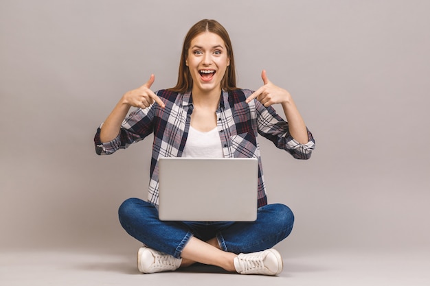 Heureuse jeune femme étonnée assise sur le sol avec les jambes croisées et utilisant un ordinateur portable