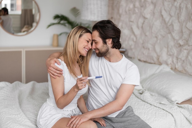 Heureuse jeune femme étreignant son mari tenant un test de grossesse positif sur le lit à la maison