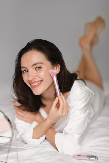 Heureuse jeune femme européenne appliquant de la poudre et du fard à joues avec un pinceau sur le visage, profitez des routines quotidiennes dans la chambre