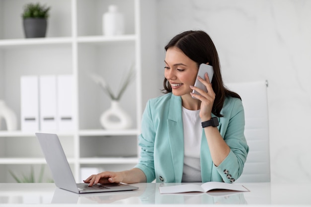 Heureuse jeune femme européenne en costume assis à table avec un ordinateur portable appelant par smartphone au bureau