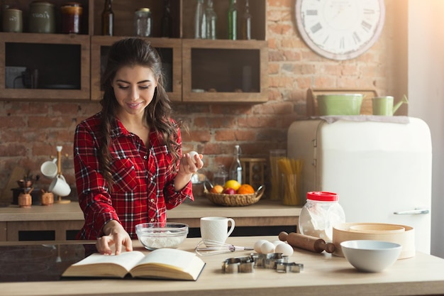 Heureuse jeune femme faisant une tarte dans une cuisine loft à la maison, à l'aide d'un livre avec une recette, espace de copie