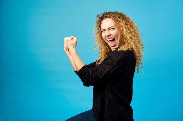 Heureuse jeune femme frisée rousse fêtée et acclamant un succès en frappant l'air avec ses poings sur le mur bleu