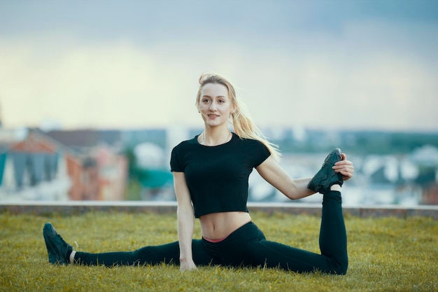 Heureuse jeune femme sur l'herbe effectue de la ficelle avec la jambe pliée sur le paysage urbain de fond