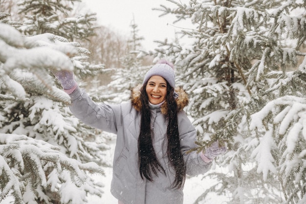 Heureuse Jeune Femme En Hiver Lors D'une Promenade Dans La Nature. Une Femme Dans Une Veste Lilas Se Tient Près Des Arbres De Noël Dans Un Parc D'hiver.