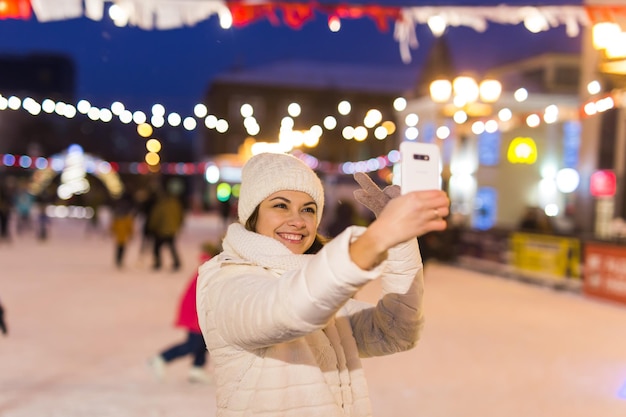 Heureuse jeune femme en hiver sur la patinoire en prenant une photo sur smartphone, selfie.