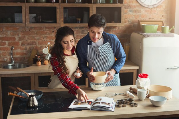 Heureuse jeune femme et homme faisant une tarte et regardant dans une recette dans une cuisine loft. Jeune famille cuisinant à la maison ensemble, espace de copie