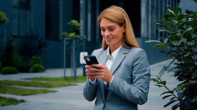 Heureuse jeune femme inspirée debout à l'extérieur tenant un téléphone portable recevant des e-mails lisant de bonnes nouvelles